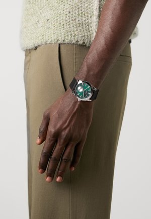 Schwarze Armbanduhr mit grünem Zifferblatt, strukturiertem Armband und silbernen Akzenten, getragen am Handgelenk einer Person in hellen Hosen.