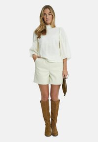 Krämfärgad blus med struktur och puffärmar, tillsammans med ljusa shorts och bruna knähöga stövlar i mocka. Håller en liten brun väska.