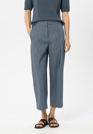 Person trägt blaue, gekürzte Hosen mit weitem Bein, blauen, kurzärmeligen Stricktop und schwarze Sandalen mit goldenen Schnallen, steht mit den Händen in den Taschen.