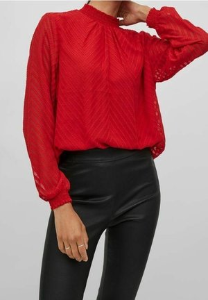 Femme portant un chemisier rouge texturé à manches longues avec col montant et un pantalon en cuir noir ajusté, posant avec une main près du cou.