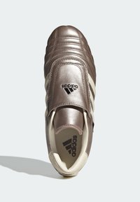 Hnědá metalická sportovní bota s texturovaným svrškem, polstrovaným límcem a třemi bílými pruhy. Na vrchu se nachází logo a má krémovou podešev.