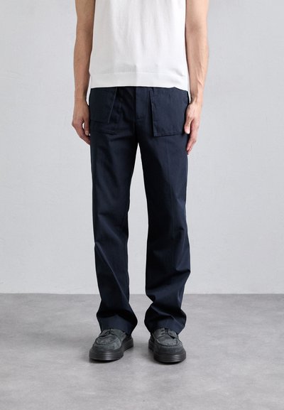Un pantalon en coton bleu marine présente une coupe ample, deux grandes poches avant et une texture lisse. Porté avec des chaussures sombres sur un sol gris.
