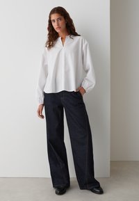 Weißes Langarmhemd mit einer Knopfleiste und einem Kragen, kombiniert mit dunkelblauen, weiten Jeans mit orangefarbenen Nähten. Schwarze Schuhe runden das Outfit ab.