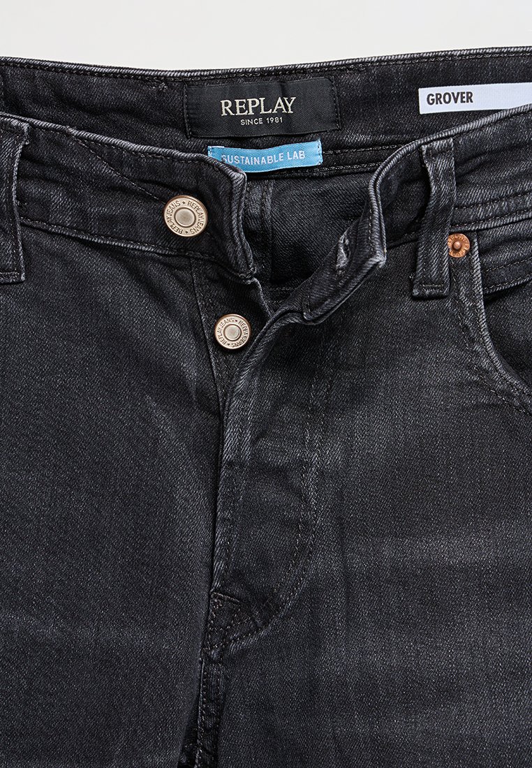 Zwarte denim jeans met knoopsluiting, metalen klinknagels en labels "Replay Since 1981," "Sustainable Lab," en "Grover" zichtbaar op de tailleband.