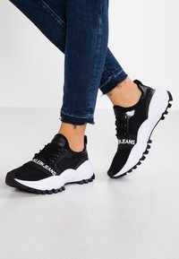 Black and white athletic shoes with a textured fabric upper, featuring a logo, chunky sole, and contrasting rubber details.