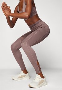 Leggings marrone a vita alta con pannelli in rete e dettagli incrociati sui polpacci, abbinati a scarpe da ginnastica bianche.