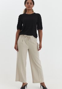 Zwarte textuurblouse met pofmouwen, gecombineerd met beige brede broek met elastische tailleband en trekkoord. Zwarte hoge muiltjes.