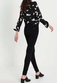 Vaqueros ajustados negros combinados con una blusa blanca y negra con estampado, de corte suelto, mangas largas y dobladillo redondeado; mules negros de tacón.