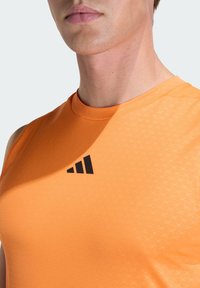 Πορτοκαλί αθλητικό φανελάκι χωρίς μανίκια με ραβδωτή υφή, διακοσμημένο με μαύρο λογότυπο Adidas στο στήθος και διακριτικά γεωμετρικά σχέδια.