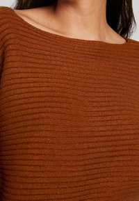 Rustfarbener Strickpullover mit geripptem Muster und weitem Ausschnitt, der horizontale Linien über den gesamten Stoff zeigt.