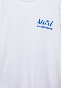T-shirt blanc avec logo "Stud Streetwear Brand" bleu imprimé sur le haut du côté gauche de la poitrine.