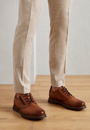 Zapatos marrones de piel texturizada con cordones y cremalleras laterales, usados con pantalones de lino beige sobre suelo de madera.