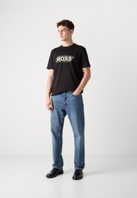 T-shirt en coton noir avec le logo "BOSS" en contour blanc texturé, associé à un jean en denim bleu clair et des chaussures noires, se tenant devant un fond uni.