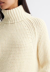 Vafflemønstret genser med høy hals i kremhvitt. Har ribbestrikket krage og drop shoulders, som viser frem et teksturert, luftig stoff.