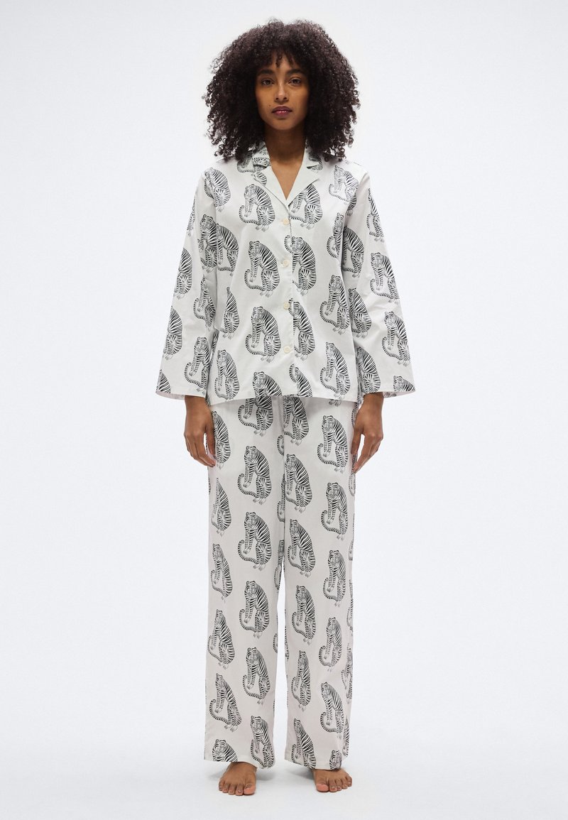 Λευκή πιτζάμα με μαύρα μοτίβα τίγρης, που περιλαμβάνει πουκάμισο με κουμπιά και φαρδιά παντελόνια, κατασκευασμένη από απαλό ύφασμα με λεία υφή.