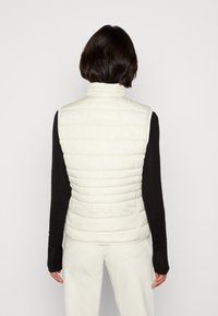 Weiße Steppweste mit horizontalen Nähten, hohem Kragen, leichtem Design und glatter Textur, getragen über einem schwarzen Langarmshirt.