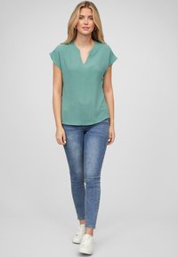 Myntegrønn kortermet topp med et subtilt teksturmønster og V-halsdetalj, kombinert med tettsittende blå jeans og hvite joggesko.