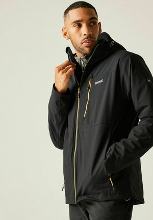 Mężczyzna poprawiający kołnierz czarnej, wodoodpornej kurtki Regatta z żółtymi suwakami, stojący na tle jednolitego jasnego tła.