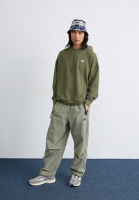 Ελαιοπράσινη κουκούλα με μπροστινή τσέπη, σε συνδυασμό με χαλαρούς φωτεινής πράσινης απόχρωσης παντελόνες. Τα αξεσουάρ περιλαμβάνουν ένα καπέλο συλλογής σε μπλε και λευκή καρώ και αθλητικά παπούτσια.