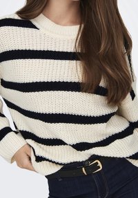 Femme portant un pull en maille crème à rayures horizontales noires, un jean bleu foncé, une ceinture noire avec une boucle dorée, et de longs cheveux bruns.