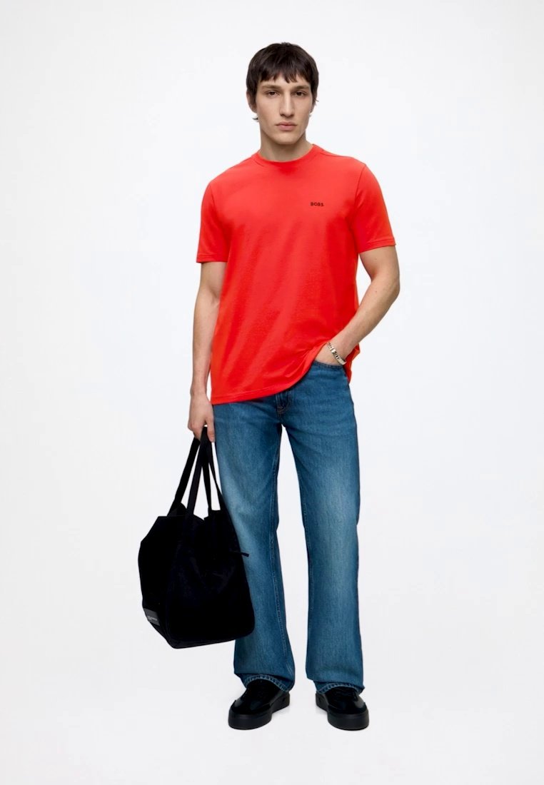 Homme portant une chemise rouge à manches courtes et un jean bleu, tenant un sac cabas noir dans la main gauche, la main droite dans la poche, debout sur fond blanc.