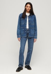 Chaqueta de denim con corte recto, dos bolsillos en el pecho, cierre de botones y costuras en contraste, combinada con jeans de denim similares y zapatos negros.