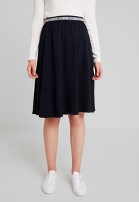 Tummansininen A-linjainen hame, jossa on joustava vyötärö ja erottuva logo-nauha. Silkkinen, kevyt kangas, polven korkeudelle ulottuva, yhdistettynä valkoisiin tennareihin.