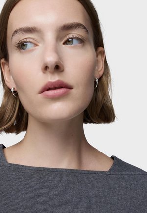 Donna con capelli corti castani che guarda di lato, indossa piccoli orecchini a cerchio argentati e una maglia grigia a costine con scollo quadrato.