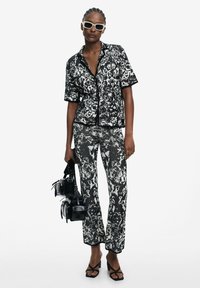 Black and white patterned short-sleeve shirt paired with matching pants; holding black bags; sunglasses; smooth fabric; tailored fit.