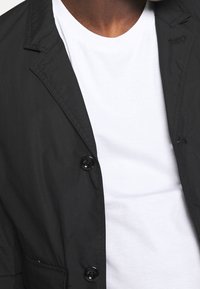 Svart, lätt jacka med två framknappar, skarpa kragar och en vänster bröstficka, bärs över en vit T-shirt med rund halsringning.