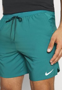 Man med petrolfärgade Nike-löparshorts med dragsko i midjan och vit swoosh-logotyp på vänster lår, hand vilande nära fickan.