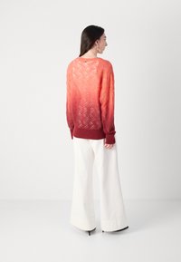 Un pull en maille présentant un dégradé du rouge à l'orange, avec des motifs en zigzag texturés. Associé à un pantalon large blanc.