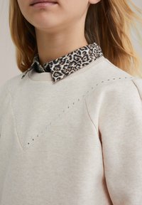 Felpa beige chiaro con un design a forma di V decorato con piccole pietre di strass. Sotto, è visibile una camicia col colletto con stampa leopardata.