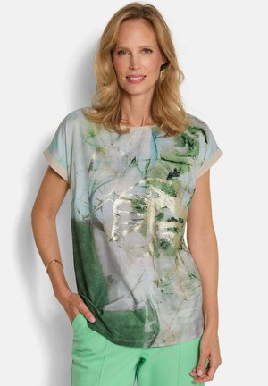 Vrouw draagt een korte mouw abstracte top met groene, grijze en gouden accenten, gecombineerd met lichtgroene broek, staand met hand in de zak.