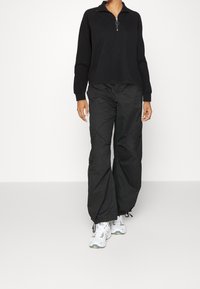 Svart zip-up sweatshirt med långa ärmar, kombinerad med löst sittande svarta cargobyxor. Bärs med vita sneakers med gröna accenter.