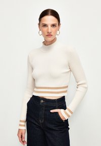 Jas beige, prążkowany sweter o krótkim kroju z wysokim kołnierzem i długimi rękawami, z brązowymi poziomymi paskami na mankietach i dolnym brzegu. Ciemne dżinsy.