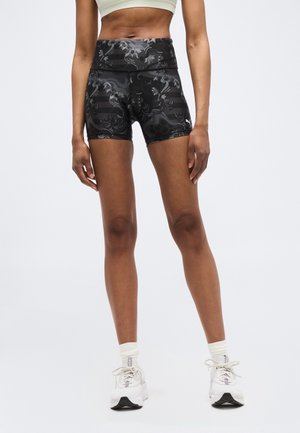 Kvinde iført sorte atletikshorts med mønster, hvid sports-bh, hvide sokker og hvide sneakers, stående mod en ensfarvet baggrund.