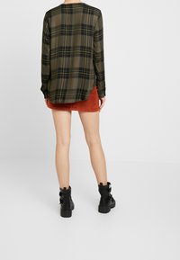 Grün karierte Langarmbluse mit schwarzen und gelben Linien, kombiniert mit einem orangefarbenen Cordrock und schwarzen Stiefeletten mit Schnallen.