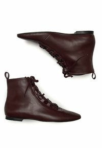 Botas de cuero marrón hasta el tobillo que cuentan con una puntera afilada, un diseño con cordones con acentos decorativos en forma de lazo y suelas planas. Textura suave en toda su superficie.