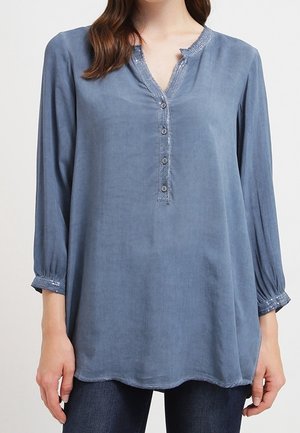 Une femme porte une tunique bleue ample à col en V, avec une patte de boutonnage et des manches trois-quarts, assortie à un jean foncé.