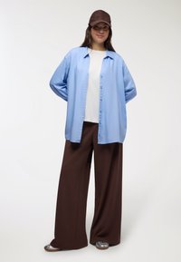 Jasnoniebieska koszula z guzikami noszona na białej koszulce, w zestawie z brązowymi spodniami o szerokich nogawkach i teksturowaną brązową czapką. Srebrne buty dopełniają całość.