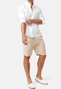 Man som rullar upp ärmarna i en vit skjorta med knappar, beige shorts och vita slip-on skor mot en enkel bakgrund.