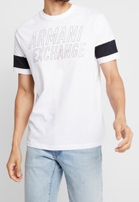 Bílá bavlněná tričko s potiskem "ARMANI EXCHANGE" v tučných šedých písmenech na přední straně, s námořnicky modrými pruhy na rukávech.