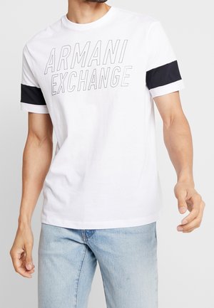 Camiseta blanca de algodón con "ARMANI EXCHANGE" impreso en letras grises en negrita en el frente, con franjas de acento azul marino en las mangas.
