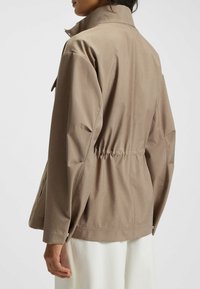 Giacca marrone chiaro con colletto alto, vita regolabile con coulisse e maniche lunghe. Presenta una texture liscia e un design dal fitting rilassato.