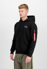 Sweatshirt preto com capuz, bolso canguru, cordões e um logótipo no peito. Possui um bolso com fecho na manga com uma etiqueta vermelha.