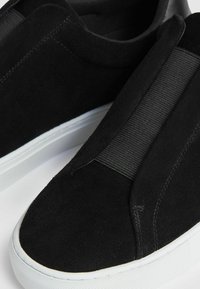Scarpe slip-on nere in suede con pannelli elastici laterali e suole in gomma bianca. Il design presenta una texture liscia e uno stile minimalista.
