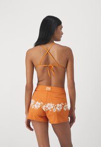 Haut de bikini orange avec des bretelles croisées et un dos à nouer, associé à un short assorti avec un motif floral blanc et un patch logo.