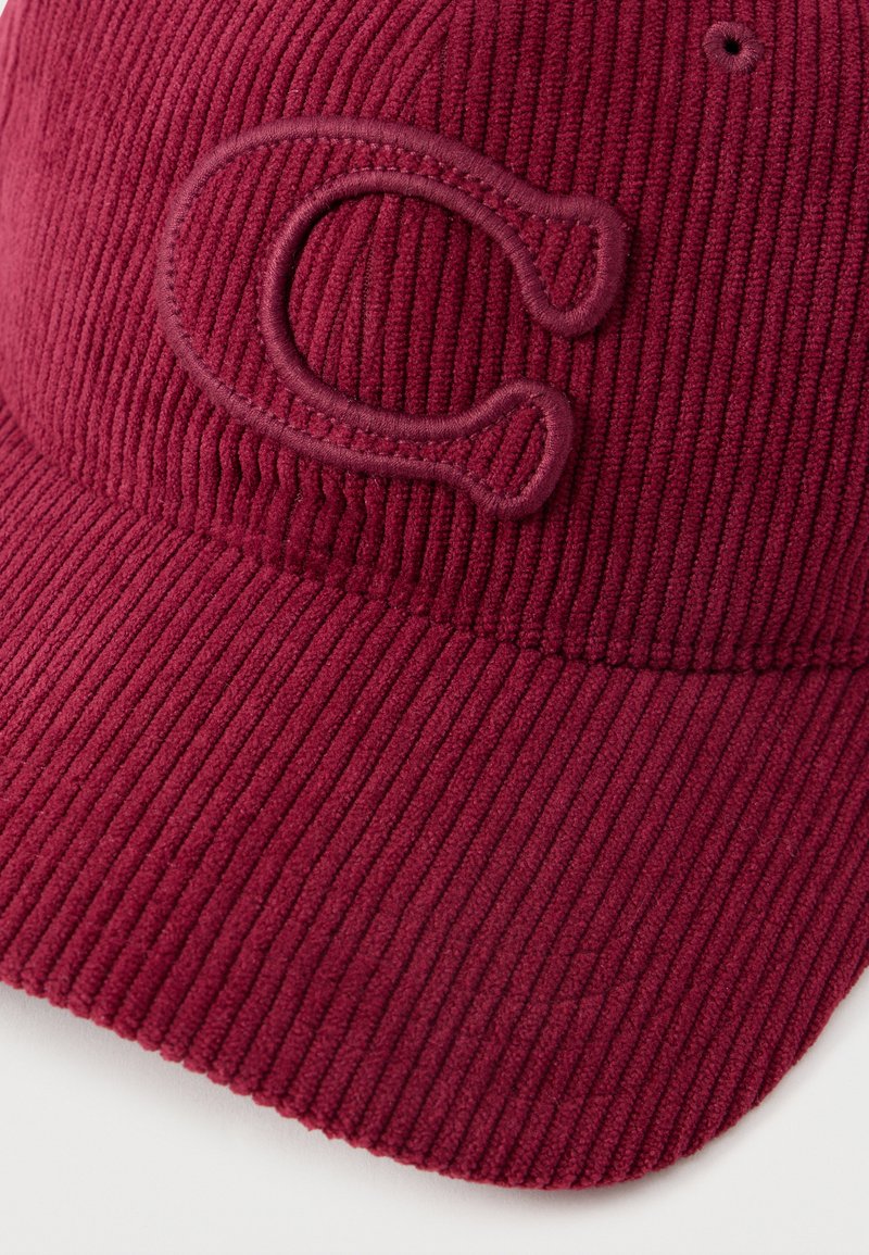 Gorra de pana color marrón con un logo "C" bordado en relieve, superficie texturizada y visera curva. El diseño es simple y moderno.