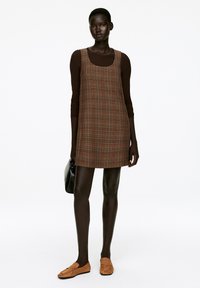 Braunes karriertes Kleid mit geradem Schnitt, über einem braunen Oberteil getragen. Kombiniert mit braunen Slippern und einer schwarzen Handtasche. Glatte Stoffstrukturen.